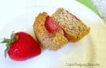 A close up of an oatmeal muffin, cut in half, on a white plate with strawberries nearby