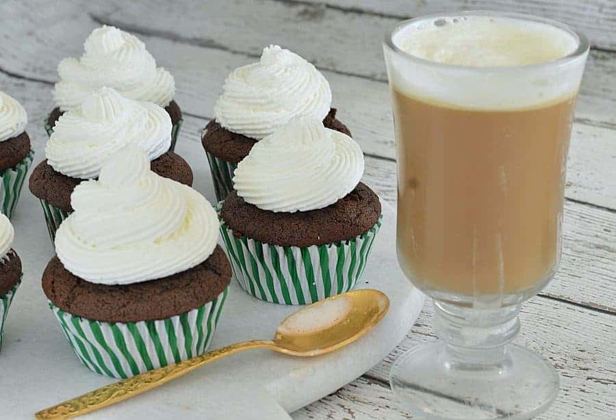 A clear glass mug of coffee next to a tray with cupcakes and a golden spoon