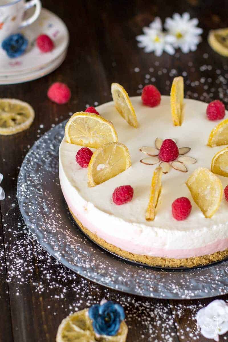 Finished no bake cheesecake with lemons, raspberries & almonds on top, sitting on a gray platter, sprinkled with powdered sugar
