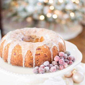 A bundt cake with glaze on top, and frosted cranberries on the side, on a white cake stand, with a lit Christmas tree in the background