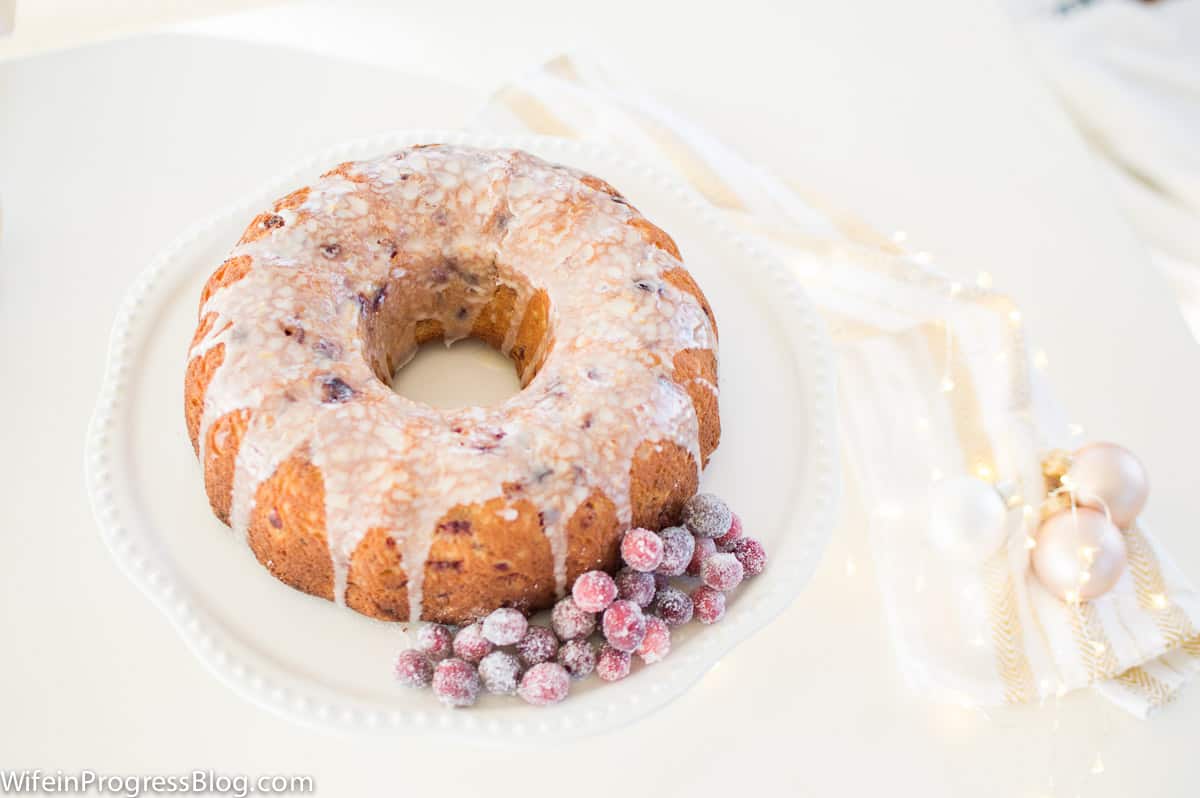 A bundt cake with glaze on top and frosted cranberries on the side, on a white cake stand