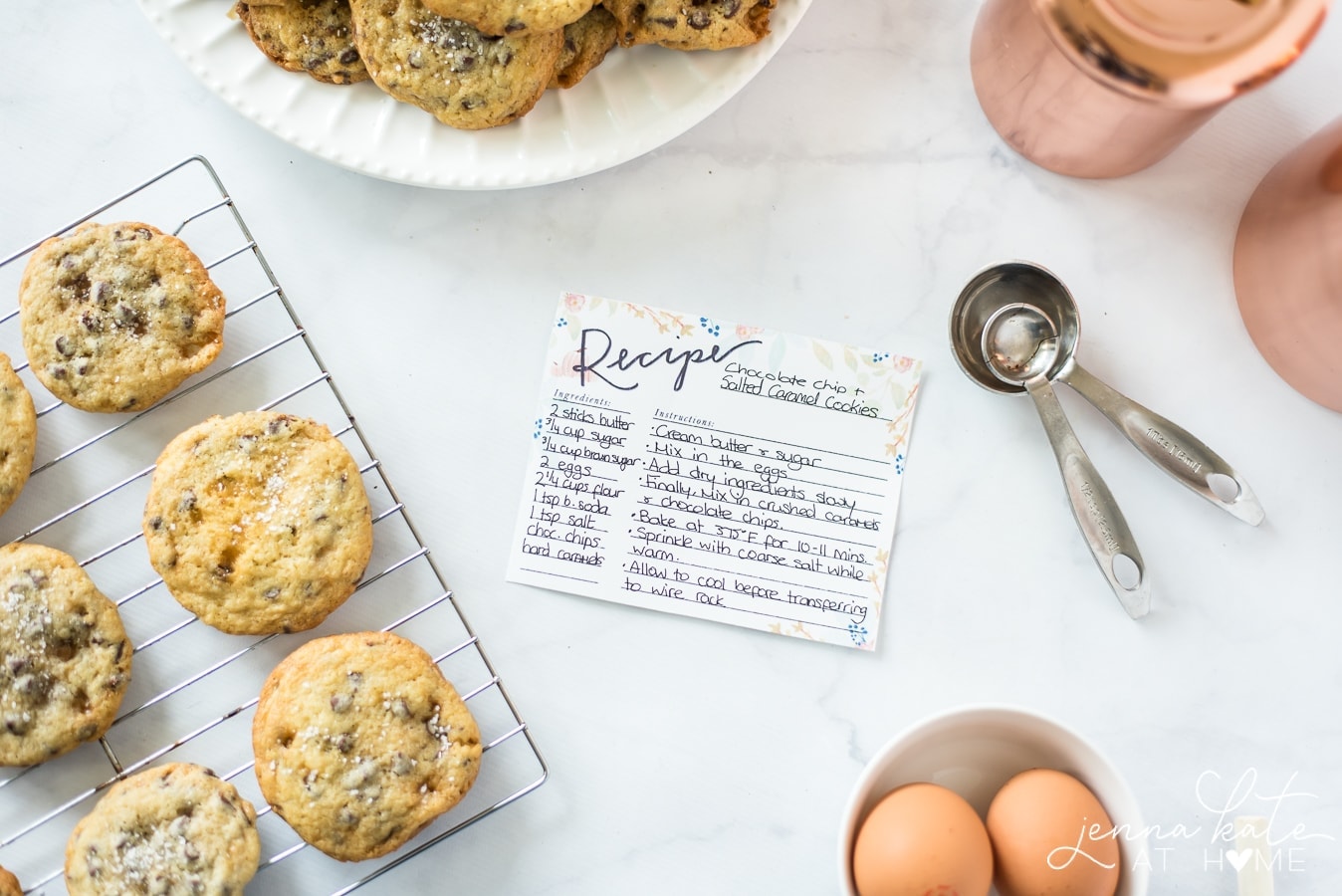 An artful display of a hand-written recipe in the center, surrounded by the edges of a plate of cookies, measuring cups and spoons, a bowl of eggs and the actual cookies on a wire cooling rack