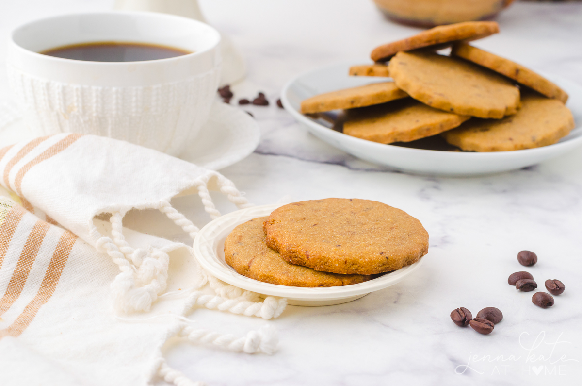 two coffee cookies on a plate next to a cup of coffee with  more cookies in the background